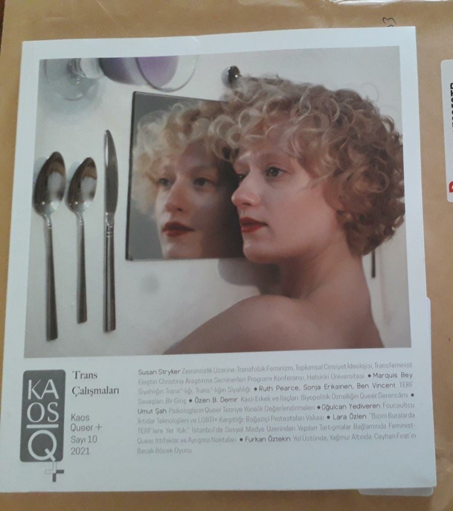 Photo of a parcel with a copy of the journal Kaos Q+ sitting on top. The journal cover features a person with blond hair and make-up lying on a table, looking blankly towards two spoons and a knife, their face reflected in a place mat.