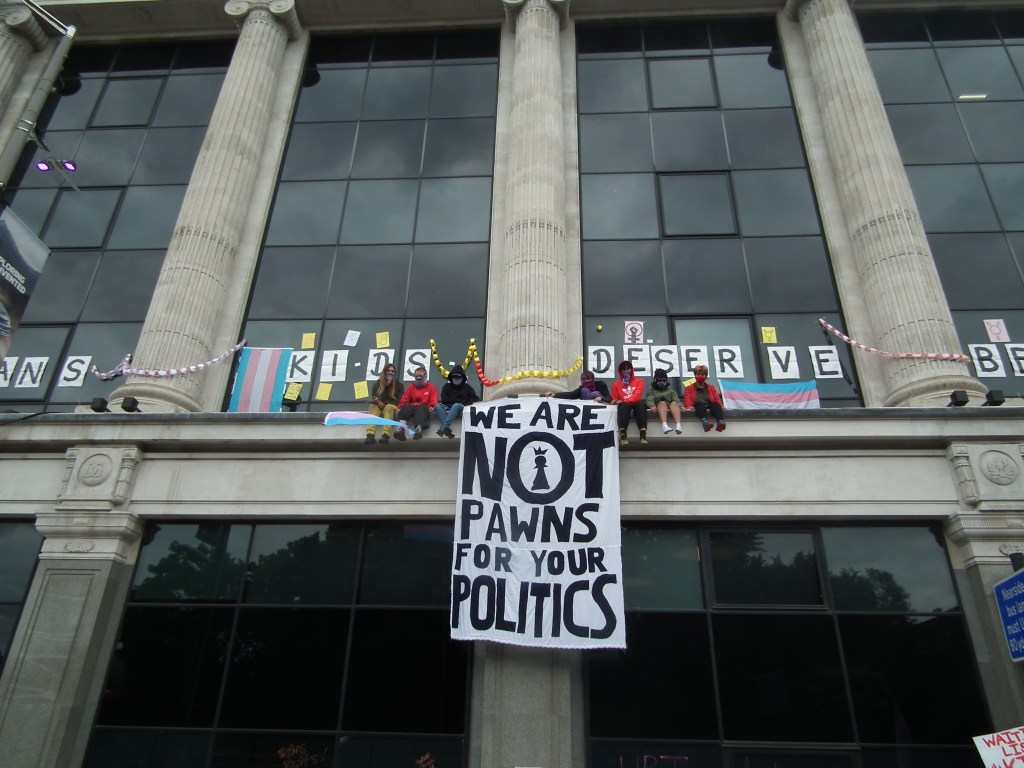 Photo of a group of people sitting on a ledge of a building, with towering pillars and glass windows behind them. They are holding a large banner which reads "We are not pawns for your politics". They have decorated with the windows of the building with trans flags, placards, and the words "trans kids deserve better".