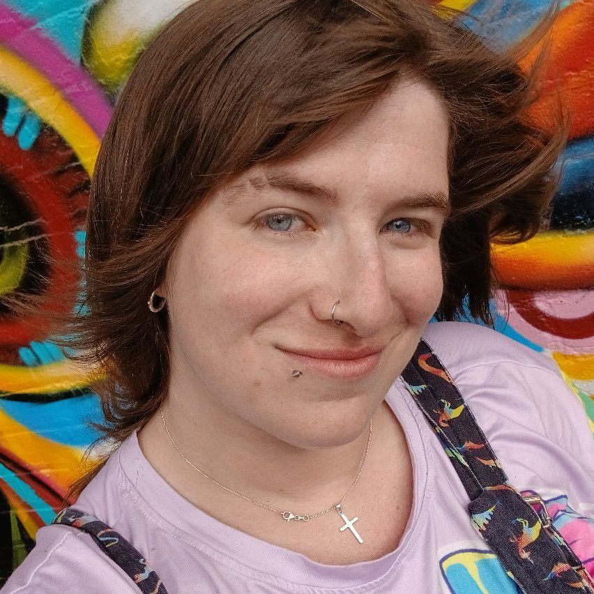 Selfie photo taken by a white woman with shoulder-length brown hair. She is wearing colourful clothes and a cross around her neck, and smiling at the camera.