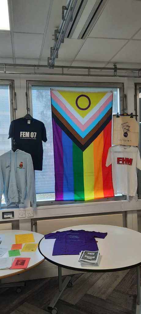Photo of WASS exhibition, with zines, flyers, t-shirts, a hoodie, a tote bag, and a progress Pride flag.